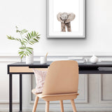 Framed print of a young African elephant on the wall behind a desk and chair. There is a pot plant on the desk and what looks like a paper weight? And a mug with a pencil in it. Some papers.